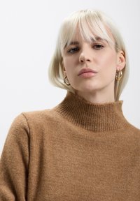 Maglione marrone, lavorato a maglia, con colletto alto a coste, texture morbida e maniche aderenti. Il modello ha i capelli biondi corti e indossa orecchini a cerchio dorati.