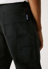 Schwarze Cargo-Hosen mit mehreren Taschen, sichtbaren Nähten und einem Gewebelabel. Das Material wirkt strapazierfähig mit einer strukturierten Oberfläche.