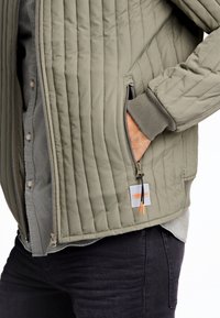 Giacca imbottita verde oliva con tasca con zip, polsini a coste e toppa con logo. Indossata sopra una camicia grigia con bottoni.