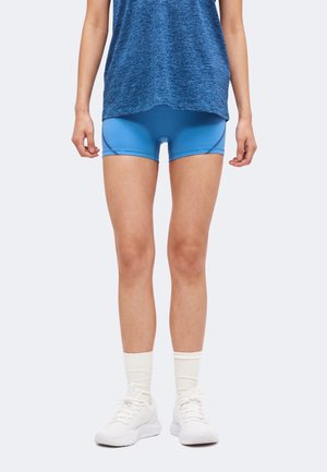 Kvinde iført blå atletikshorts, en blå meleret top, hvide ankelsokker og hvide løbesko, stående på en ensartet lys baggrund.