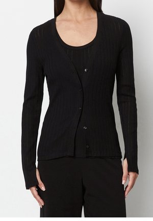 Femme portant un cardigan noir ajusté à manches longues en maille côtelée, boutonné au milieu, sur un débardeur noir assorti et un pantalon noir.