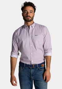 Lichtroze button-up shirt met marineblauwe stippen, lange mouwen opgerold, en een logo op de borst; gecombineerd met denim jeans en een geweven blauwe riem.