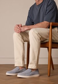 Blauwe instappers van gestructureerde stof met een witte rubberen zool, gedragen met beige broek, zittend op een houten stoel.