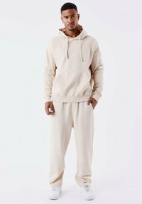 Felpa beige con cappuccio e pantaloni della tuta abbinati in morbido tessuto, caratterizzati da una tasca a marsupio e dettagli con logo sulla parte anteriore, con caviglie strette.
