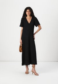 Black maxi dress with short sleeves, deep V-neck, scalloped edges, and flowy fabric. Model holds a brown handbag, wearing strappy heels.