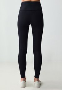 Leggings neri a vita alta realizzati in tessuto elasticizzato, con una texture liscia e un design aderente, abbinati a sneakers bianche.