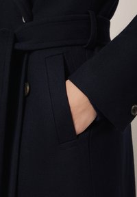 Szczegół marynarskiego płaszcza wełnianego z ręką w bocznym kieszeni, z paskiem i ozdobnym guzikiem przy mankiecie. Miękka tekstura i dopasowany krój.