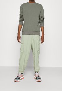 Felpa in cotone grigia, pantaloni cargo verde chiaro con tasche e sneaker grigie con accenti arancioni. Stile casual, vestibilità rilassata e tessuto strutturato.