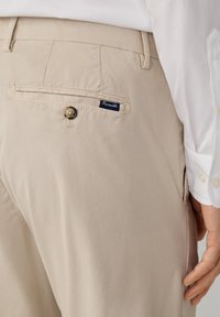 Pantaloni beige con un design dritto, dotati di una tasca posteriore con chiusura a bottone e un piccolo etichetta con il marchio. Tessuto dalla texture liscia.