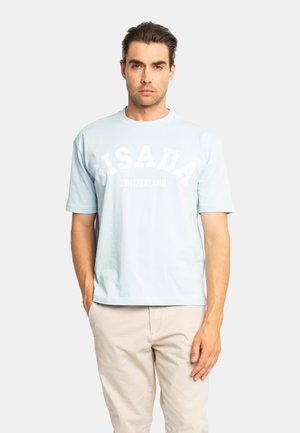 Mann mit zurückgekämmtem dunklem Haar trägt hellblaues kurzärmeliges T-Shirt mit Text "Bisada Switzerland" und beige Hose, steht vor einem einfarbigen Hintergrund.