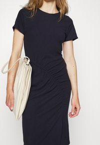 Marinblå klänning med kort ärm och rynkade sidodetaljer, tillverkad av mjukt tyg. Sammanfogad med en stor beige kuvertväska.