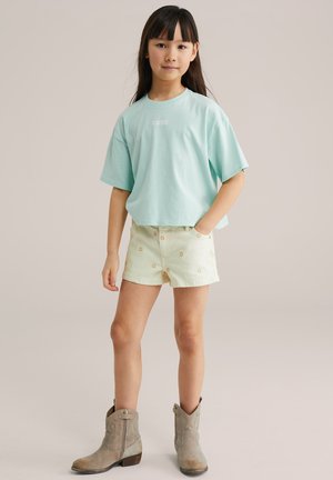 Noor tüdruk seisab ühe käega taskus, kandes lahtist helesinist t-särki, kreemikaid lühikesi pükse, millel on väikesed lillemustrid, ning beeže pahkluu saapaid.