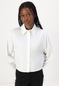 Hvit cropped knapp-up skjorte med lange ermer, stiv krage og en jevn tekstur. Skjorten har et enkelt, klassisk design.