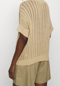 Valkoinen neulepaita, jossa on löysä istuvuus, lyhyet hihat ja teksturoitu avoin kuvio. Yhdistetty beigeihin shortseihin kerroksellisen ilmeen saavuttamiseksi.