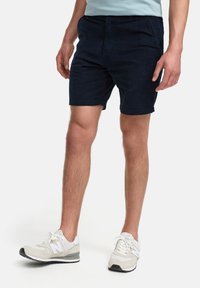Marinblå bomullsshorts med platt framsida, två sidofickor och uppvikt fåll, tillsammans med vita sportskor med grå detaljer.