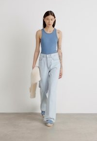 Lichtblauw tanktop, hoogwaisted licht denim jeans en blauwe sneakers. Een model loopt op een neutrale achtergrond terwijl ze een crèmekleurige cardigan vasthoudt.