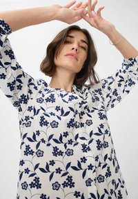 Hvid bluse med marineblåt blomsterbroderi, der har trekvartærmer, en rund halsudskæring og knapper langs fronten.