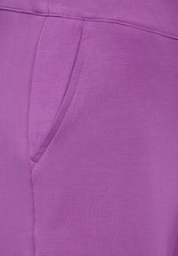 Tissu violet avec une texture lisse, présentant une poche latérale et des détails de couture. Le matériau semble léger et flexible.