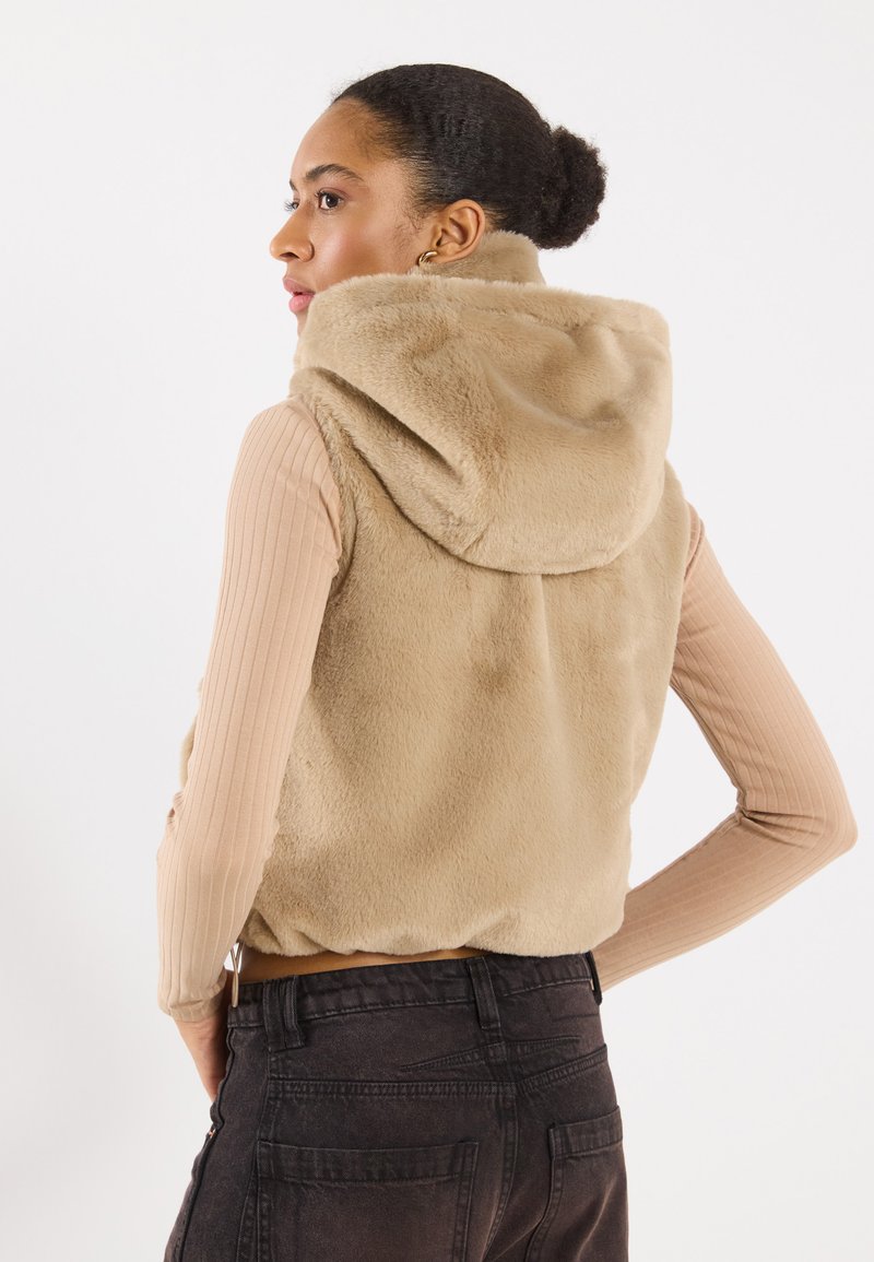 Beige nepbont vest met een capuchon, voorzien van een zachte textuur. Een geribbelde lange mouwtop eronder en een donkere spijkerbroek maken de outfit compleet.