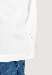 Camicia bianca in cotone a maniche lunghe con orlo dritto, caratterizzata da un piccolo logo sulla cucitura laterale sinistra, abbinata a jeans in denim blu.