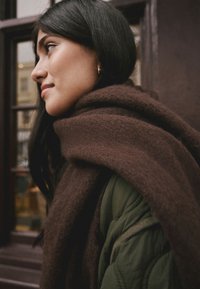 Brown wool scarf draped over an olive green quilted jacket. Close-up of a face with long black hair, featuring a simple gold earring.