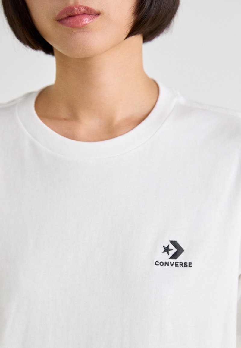 Camiseta de algodón blanca con cuello redondo y un logo negro de Converse con un diseño de estrella y flecha en el lado izquierdo del pecho. Textura suave.