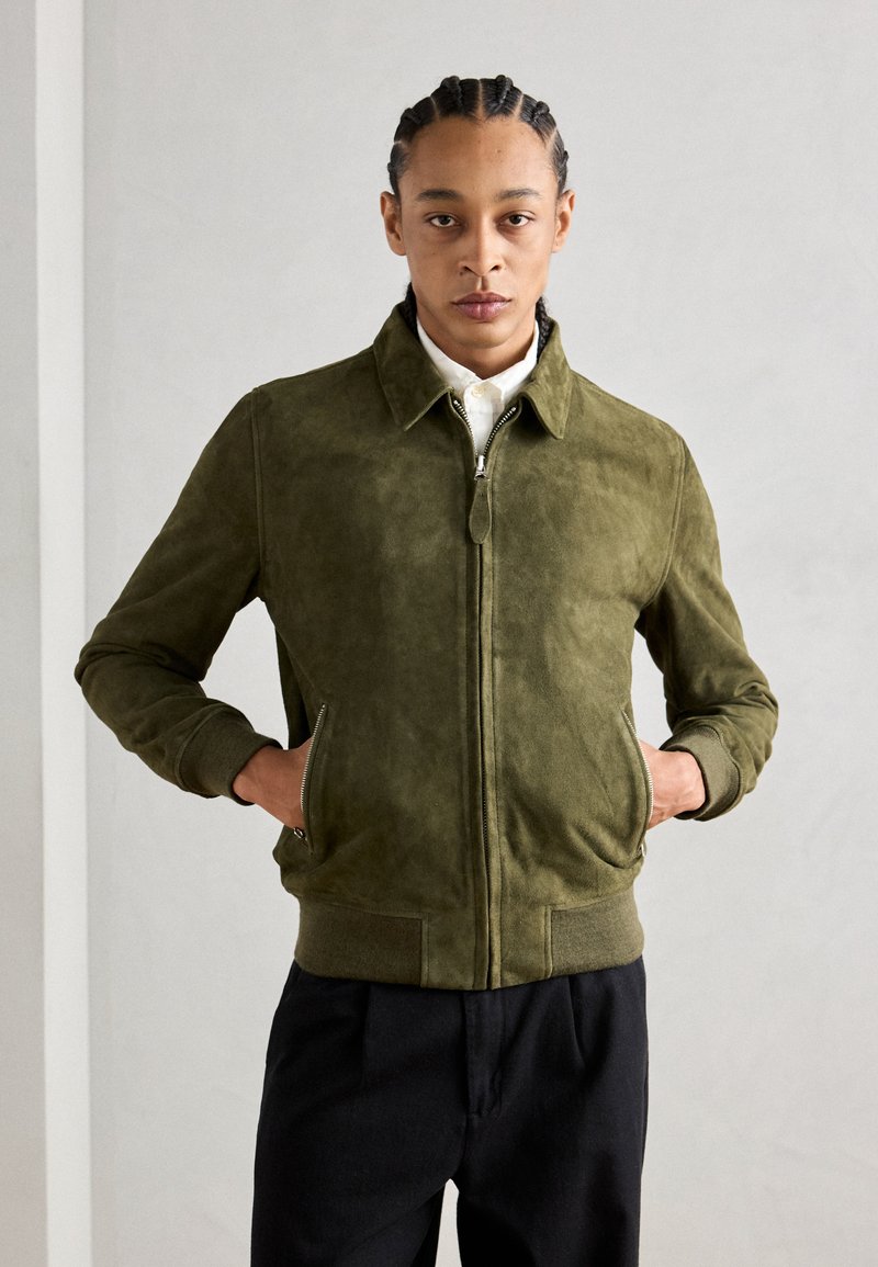 Giacca bomber in suede verde oliva con colletto, chiusura con zip e tasche laterali. Polsi e orlo a coste; abbinata a una camicia bianca.