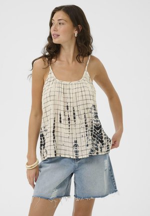 Žena v bezrukávovom krémovo-čiernom top s mriežkovým vzorom a džínsových kraťasoch, pozerajúca sa do strany, na neutrálnej pozadí.