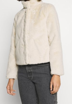 Personne portant une veste duveteuse couleur crème à manches longues, associée à un jean taille haute gris foncé sur un fond uni.