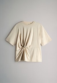 Beige short-sleeve t-shirt with a twisted knot detail at the waist, made from soft fabric. Crew neckline and relaxed fit with smooth texture.