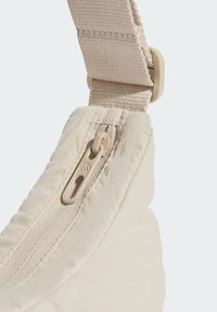 Bolso beige claro con un material suave y texturizado. Cuenta con una cremallera visible con una lengüeta y una correa ajustable de tela.