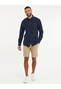 Camisa de botones azul marino en una tela texturizada, combinada con pantalones cortos beige y zapatillas blancas. Presenta un pequeño logo en el pecho, diseño casual.