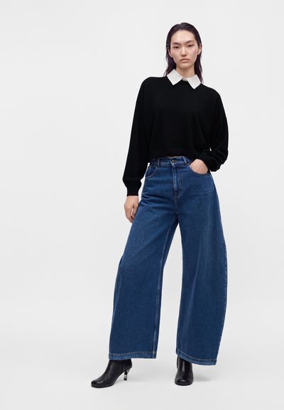 Jeune femme aux longs cheveux foncés portant un pull noir par-dessus une chemise blanche à col, un jean bleu à jambes larges et des bottines noires.
