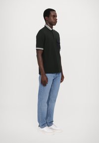 Mørk grønn polo skjorte med hvitstripet krage, lommepose på venstre bryst, kombinert med lyseblå jeans og hvite sneakers. Sideprofil.