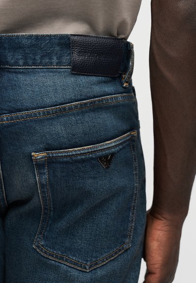 Κοντινό πλάνο της πίσω τσέπης και της ζώνης μέσης ενός μπλε τζιν παντελονιού που φοράει άτομο με σκούρο δέρμα και μπεζ πουκάμισο μαζεμένο μέσα.