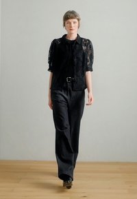 Giacca corta in pizzo nero con maniche corte sopra una maglia nera, abbinata a pantaloni neri ampi e scarpe nere, su uno sfondo neutro.