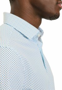 Hellblaues Hemd mit einem Kragen und einem Muster aus kleinen blauen Punkten. Der Stoff wirkt glatt und der Schnitt ist maßgeschneidert.