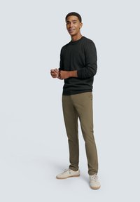 Pull à col rond gris foncé, pantalon vert olive et baskets claires. Le modèle se tient debout, les mains jointes, souriant devant un fond uni.