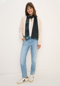 Zwarte sjaal met groene geometrische patronen, lichtgewicht crèmekleurige jas, witte top, lichtblauwe jeans en witte sneakers met zwarte accenten.