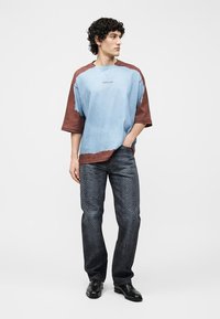 Νέος άνδρας που στέκεται με το ένα χέρι στην τσέπη, φοράει μπλε και καφέ υπερμεγέθη μπλουζάκι, σκούρο παντελόνι με σχέδια και μαύρα επίσημα παπούτσια, κοιτάζει προς το πλάι.