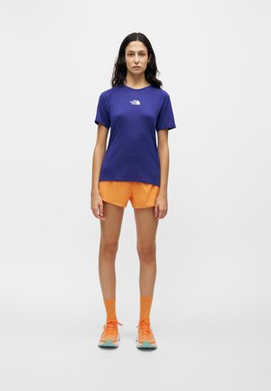 Kvinde stående iført en blå kortærmet skjorte, orange atletikshorts, orange sokker og orange løbesko på en ensfarvet hvid baggrund.
