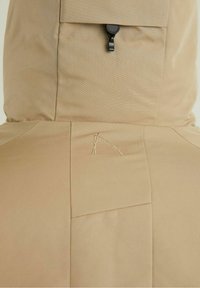 Lekka beżowa odzież wierzchnia wykonana z tkaniny o fakturze. Charakterystyczne detale to czarny zamek błyskawiczny oraz mały haft w kolorze złotym.