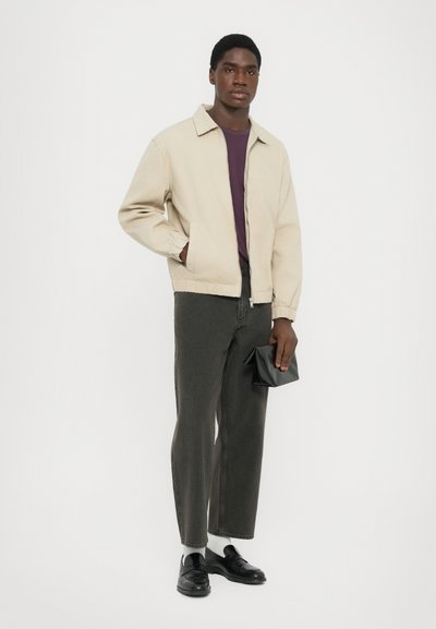Man i beige jacka, lila skjorta, mörkgrå jeans, vita strumpor, svarta loafers, håller en liten svart clutch mot vit bakgrund.