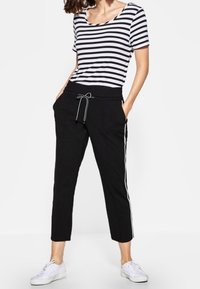 Czarne sportowe spodnie z elastycznym pasem i białymi paskami po bokach, połączone z bluzką w czarno-białe paski, noszone z białymi sneakersami.