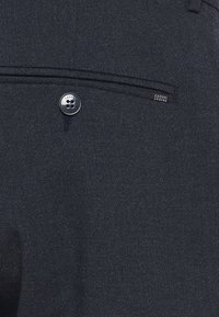 Mørkegrå dressbukser laget av teksturert stoff. Har en baklomme med en sølvknapp og et merke som sier "CASUAL FRIDAY."