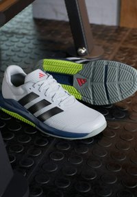 Zapatillas deportivas blancas con rayas negras, parte superior de malla texturizada y detalle de suela verde vibrante. La suela presenta un diseño de patrón para mayor agarre.