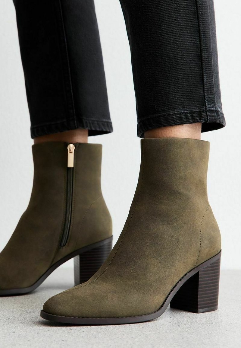 Bottes en daim vert olive montant à la cheville avec fermeture éclair latérale et talons blocs, portées avec un pantalon noir raccourci sur un sol clair.