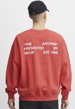 Felpa rossa con testo bianco sul retro: "L'ARCHIVIO" "PRESENTATO DA SOLID EST. 1999." Collo a giro, vestibilità ampia, polsini e orlo a coste.