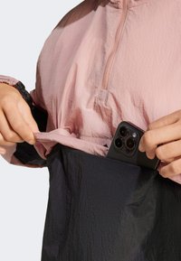 Lättviktig rosa och svart jacka med dragkedja vid halsen, texturerad tyg och en framficka där en smartphone är delvis synlig.