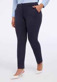 Pantaloni bleumarin, croiți dintr-un material elastic. Prezintă un design cu fața netedă, buzunare laterale și un croi ajustat, asortați cu tocuri albe.
